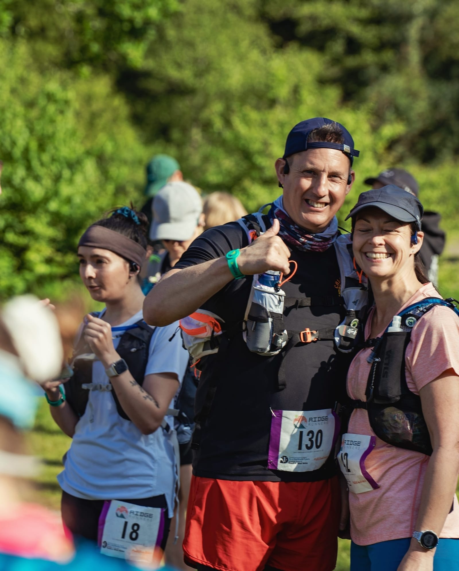 Two smiling runners wearing race bibs and hydration packs pose together outdoors. The man gives a thumbs up while the woman smiles. Other participants stand in the background, surrounded by greenery.
