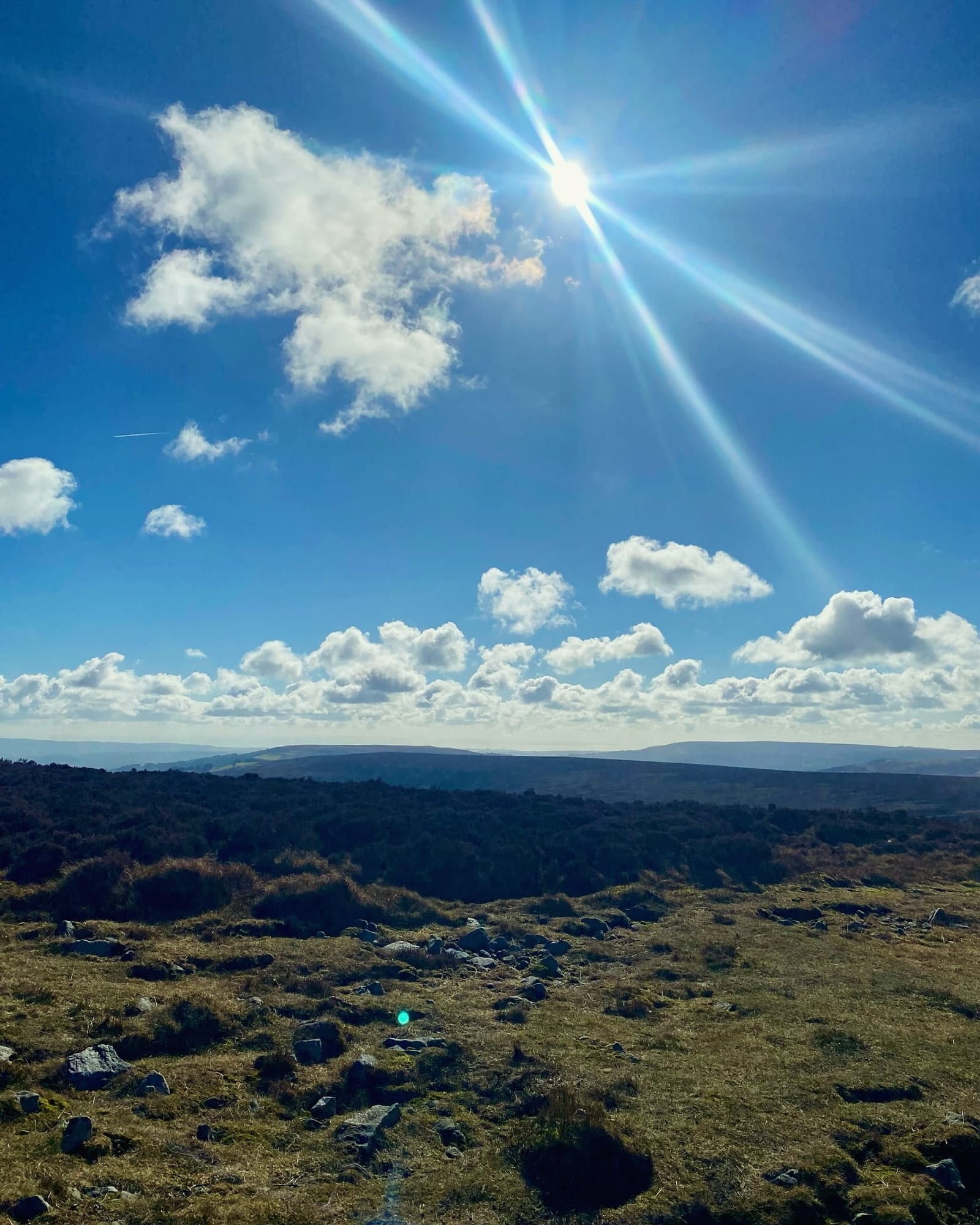 Bright sun shining through scattered clouds over a grassy, rocky landscape with hills in the distance, creating a lens flare across the clear blue sky.
