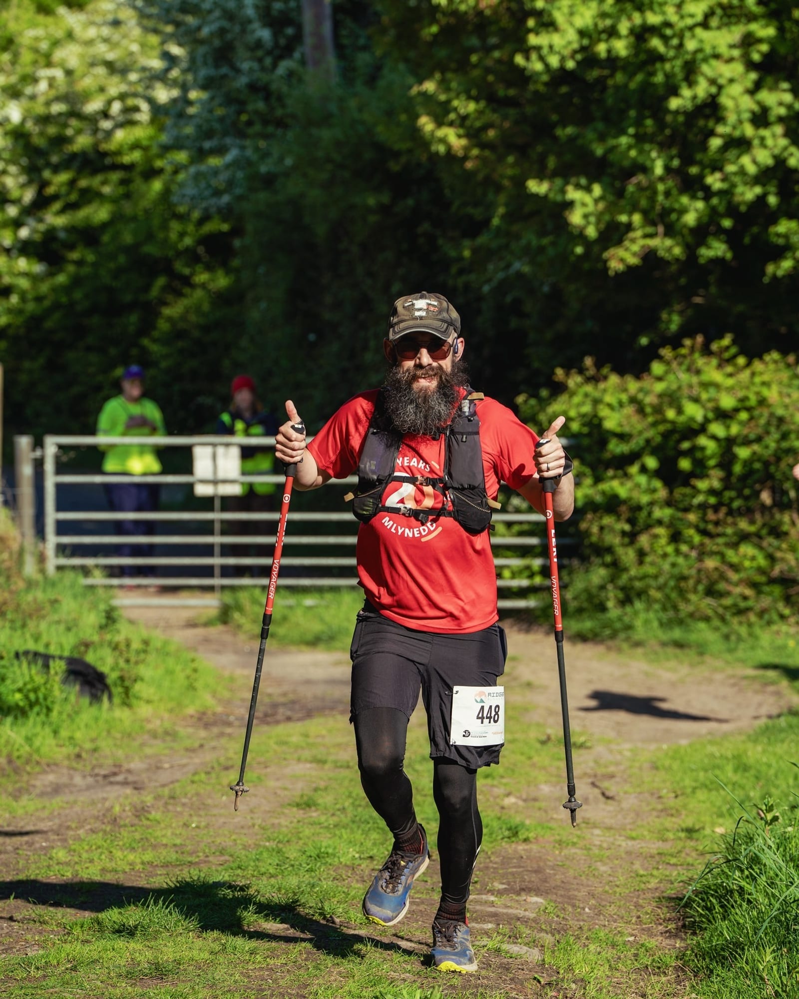 A bearded man wearing sunglasses, a red shirt, and a race bib is running on a dirt path with trekking poles, giving a double thumbs-up. Sunlight filters through greenery in the background.