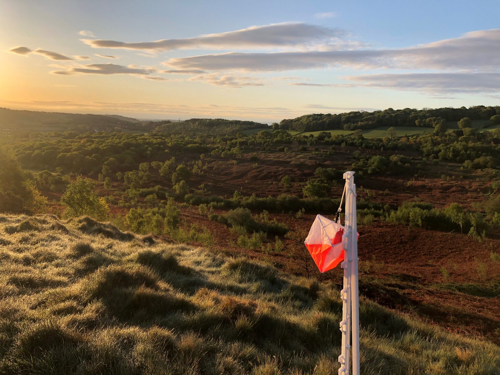 A red and white orienteering flag is placed on a grassy hilltop at sunrise. The landscape below features rolling hills covered in green vegetation and trees, with a mix of sunlight and shadows. The sky is partly cloudy with soft colors from the rising sun.