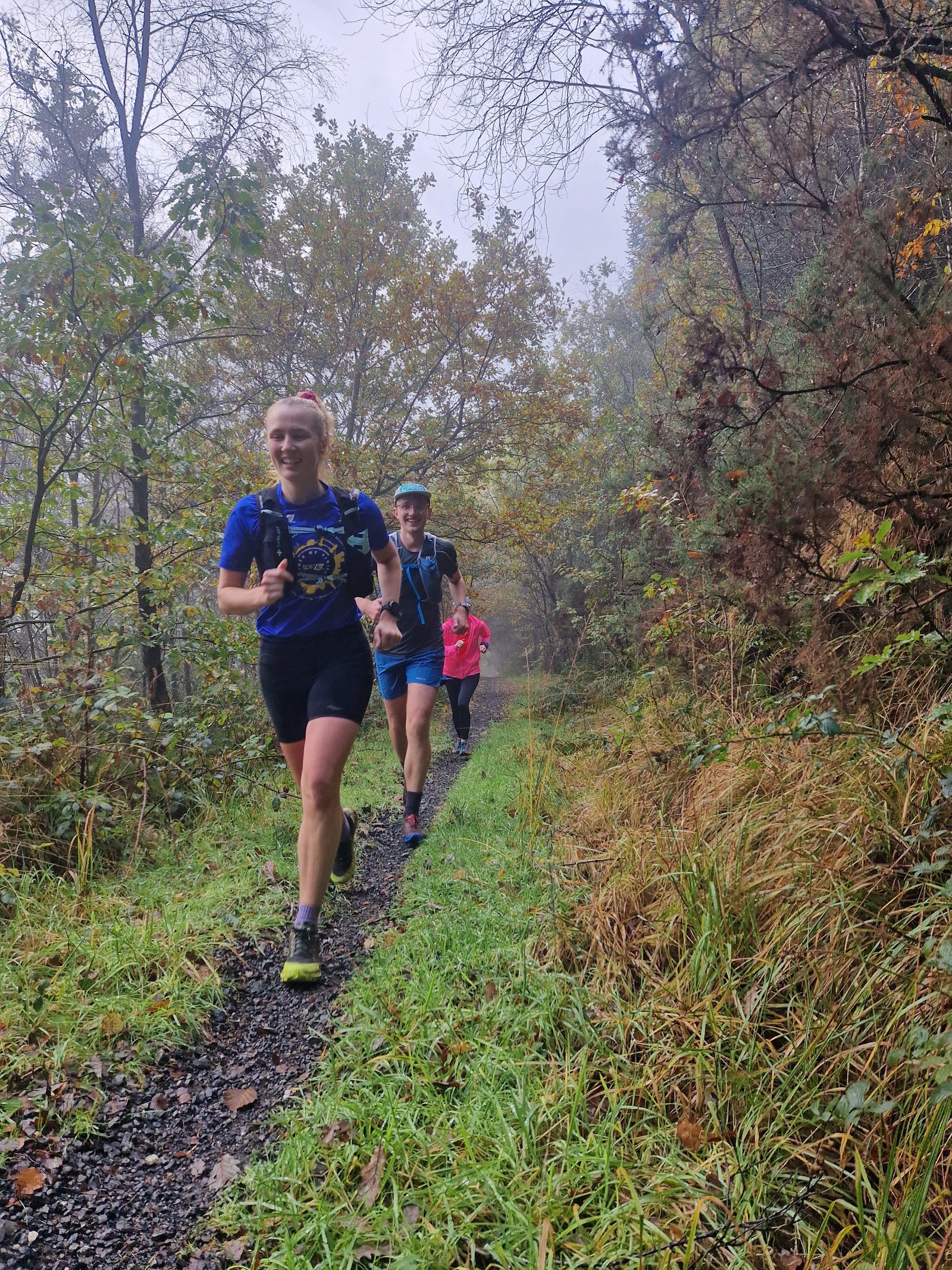 Three trail runners on a narrow forest path. The leading runner wears a blue shirt and black shorts, followed by a runner in a light blue cap. The third runner is further back and dressed in red. The surrounding trees are lush, and the ground is slightly misty.