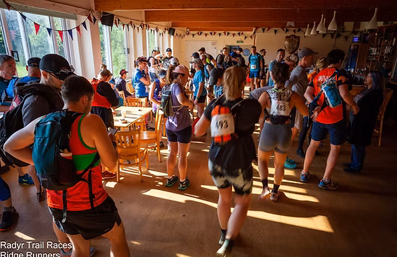 A group of trail runners wearing race bibs and backpacks gather inside a sunlit room, chatting and preparing before an event. Tables and chairs are scattered around, and festive bunting decorates the ceiling.