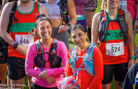 Two smiling women wearing race bibs and hydration packs stand among other runners at an outdoor trail race event. Sunlight highlights their faces, and participants are dressed in colorful athletic gear.