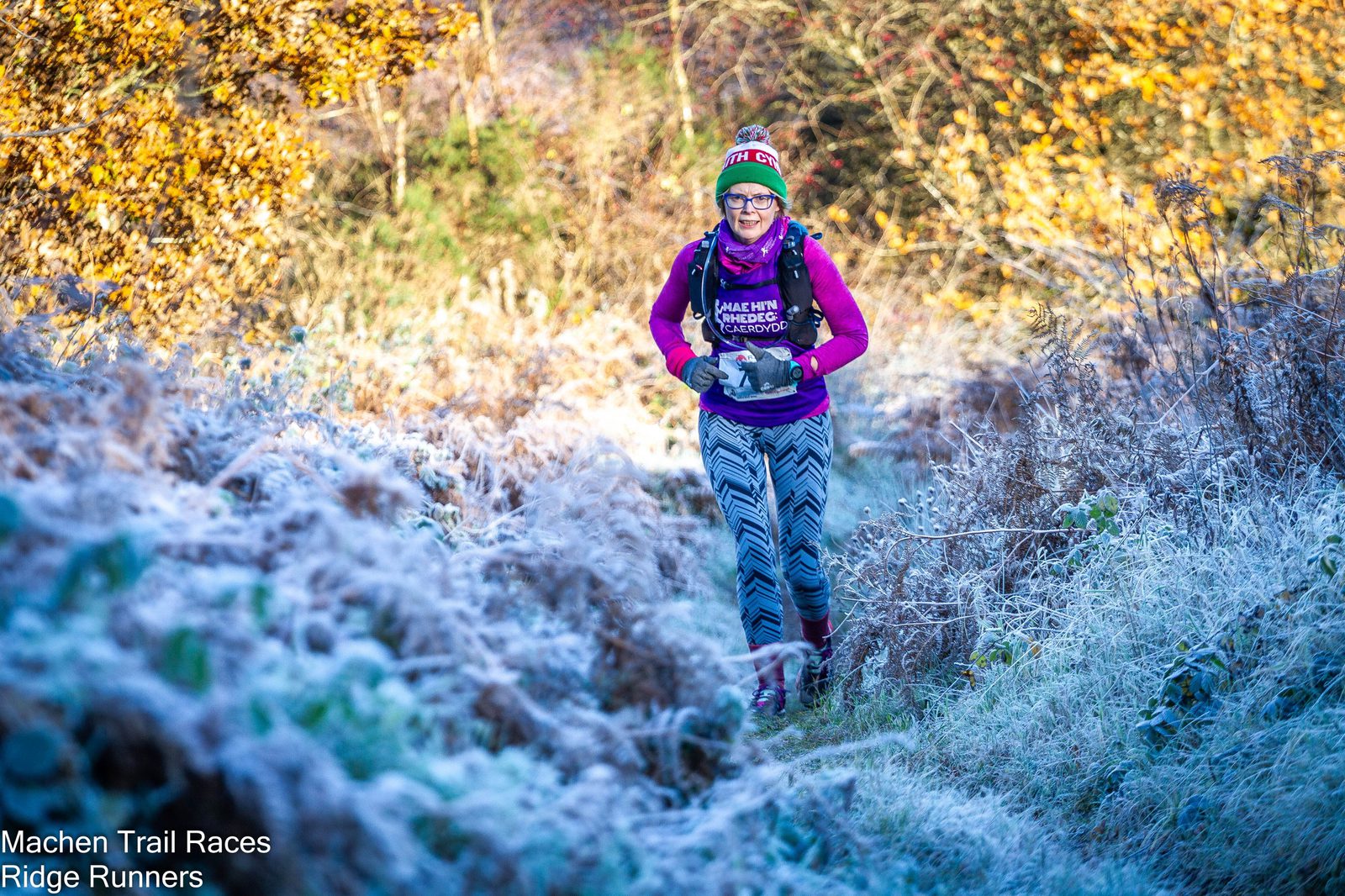 A runner dressed in a colorful beanie, glasses, purple top, and patterned leggings jogs along a frosty, narrow trail surrounded by autumn foliage. The person is participating in Machen Trail Races, as noted by the text in the image: "Machen Trail Races Ridge Runners.