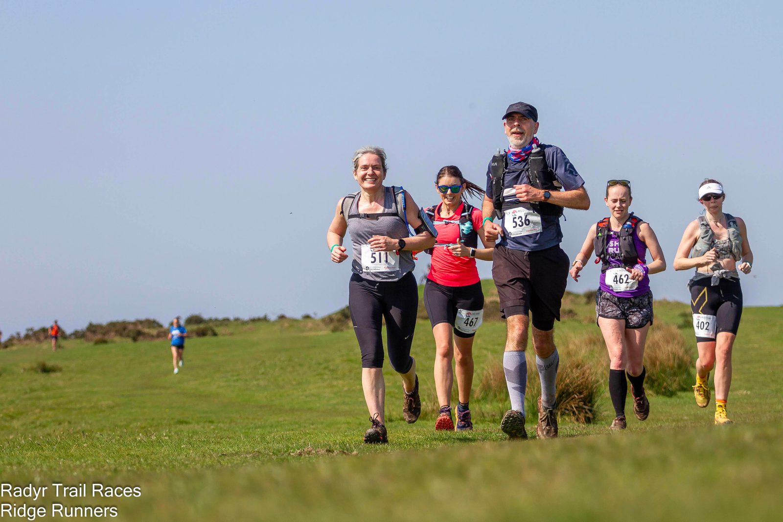 A group of six runners, wearing race bibs, run along a grassy trail under a clear blue sky. In the foreground, two women and four men are visible, with varying running gear. The background features a distant single runner and a vast green landscape. Text reads "Radyr Trail Races, Ridge Runners.
