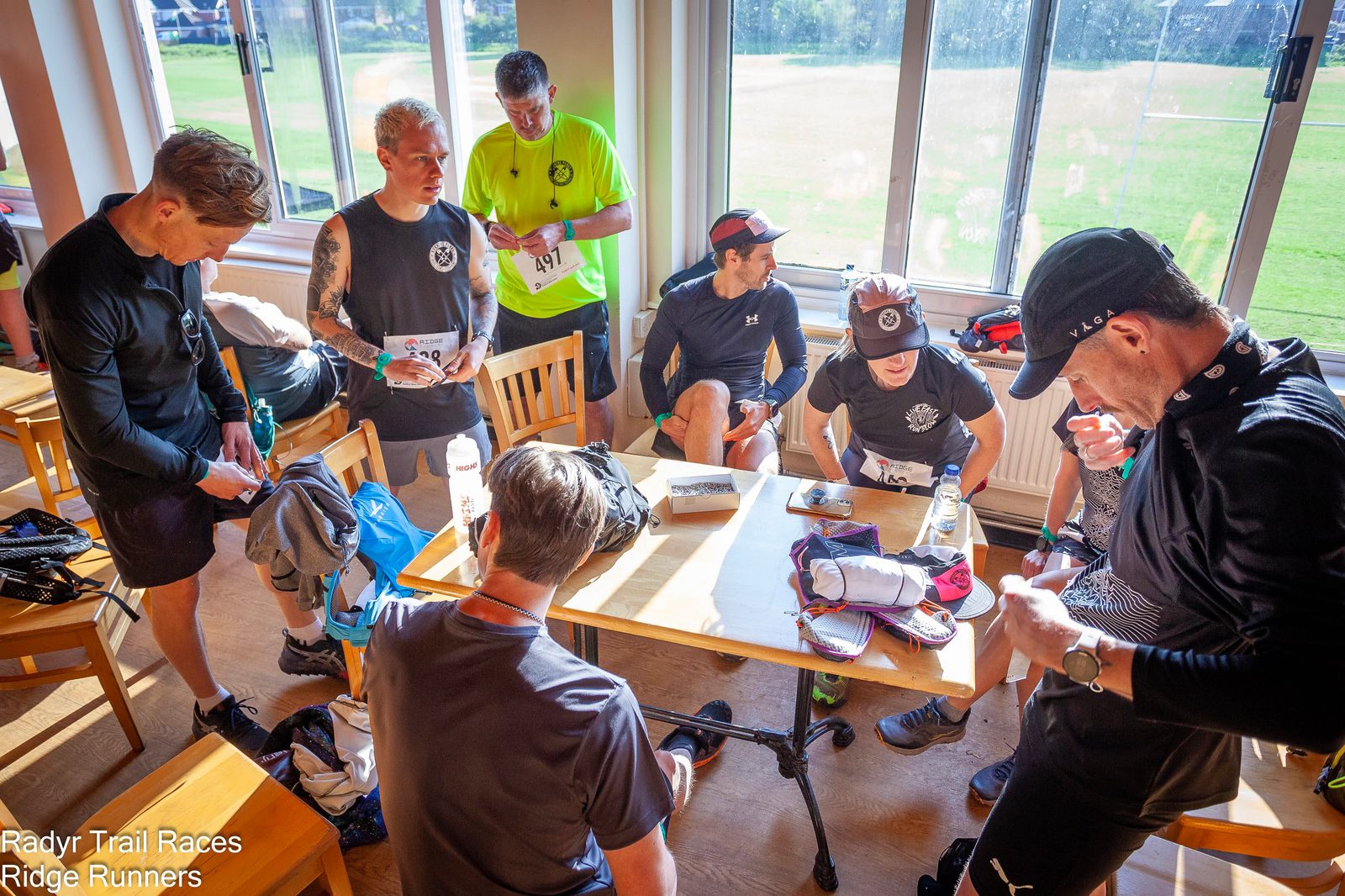 A group of runners in athletic gear gather around a table in a sunlit room, preparing for the Radyr Trail Races. Some are standing while others are seated, engaging in various tasks like checking their race numbers or gear. "Radyr Trail Races Ridge Runners" is printed on the photo.