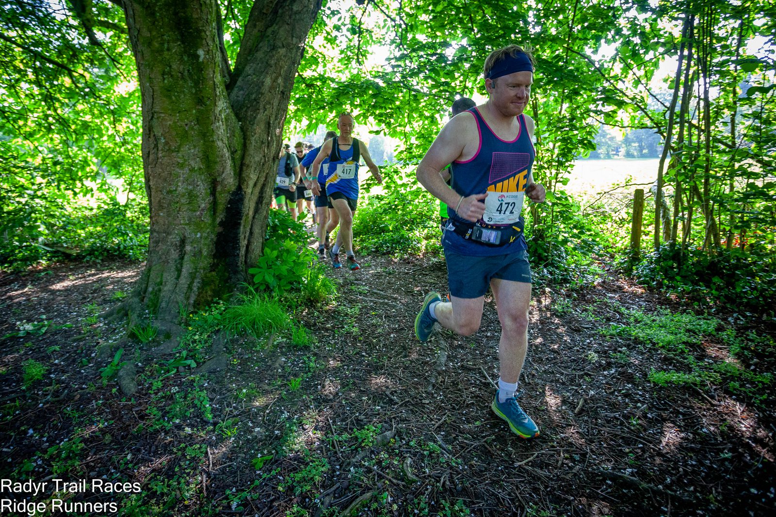 Runners participating in a trail race pass through a wooded area on a narrow path. The front runner wears a headband, sleeveless shirt, and shorts with the number 472 bib. Other runners follow closely behind. The environment is lush with greenery.