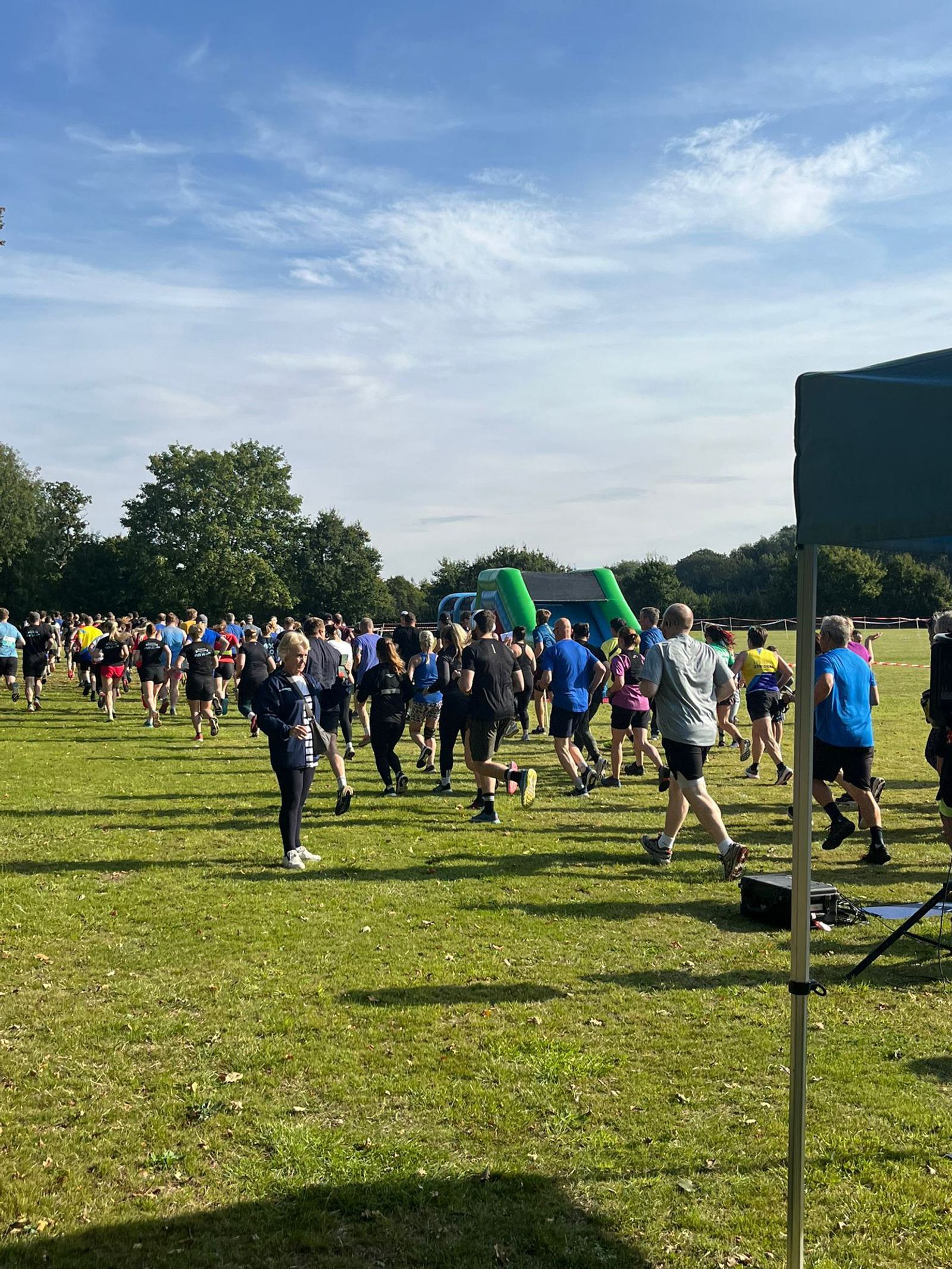 A large group of people participating in an outdoor run or race on a grassy field under a blue sky. Runners wear athletic clothing in various colors, and there is a green inflatable obstacle or marker in the background.