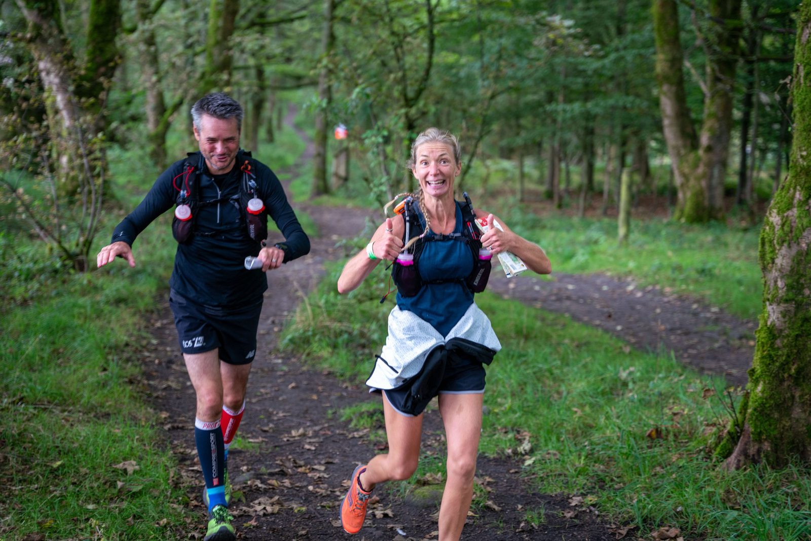 Two runners are on a forest trail, both wearing hydration packs and athletic attire. The woman in front is smiling and giving a thumbs-up, while the man behind her is focused on running. The path is surrounded by lush green trees and grass.