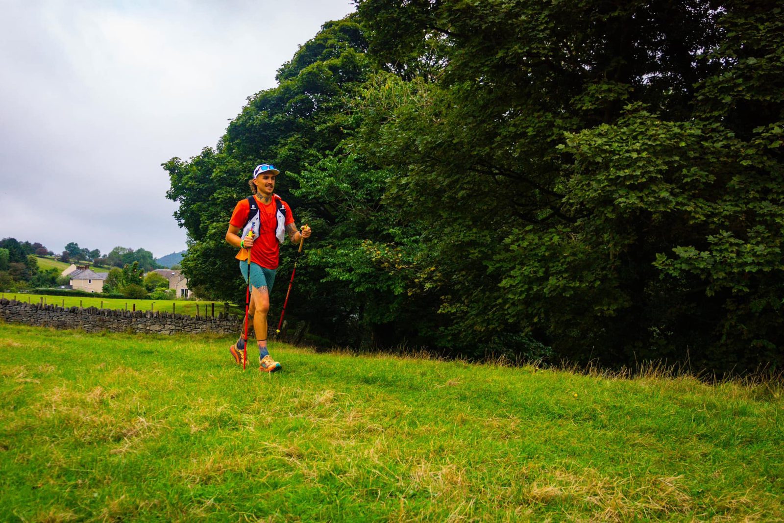 A person in outdoor gear, including a red shirt and backpack, is hiking with trekking poles across a grassy field. They appear to be enjoying the hike, with greenery and a few houses visible in the background. The sky is overcast.