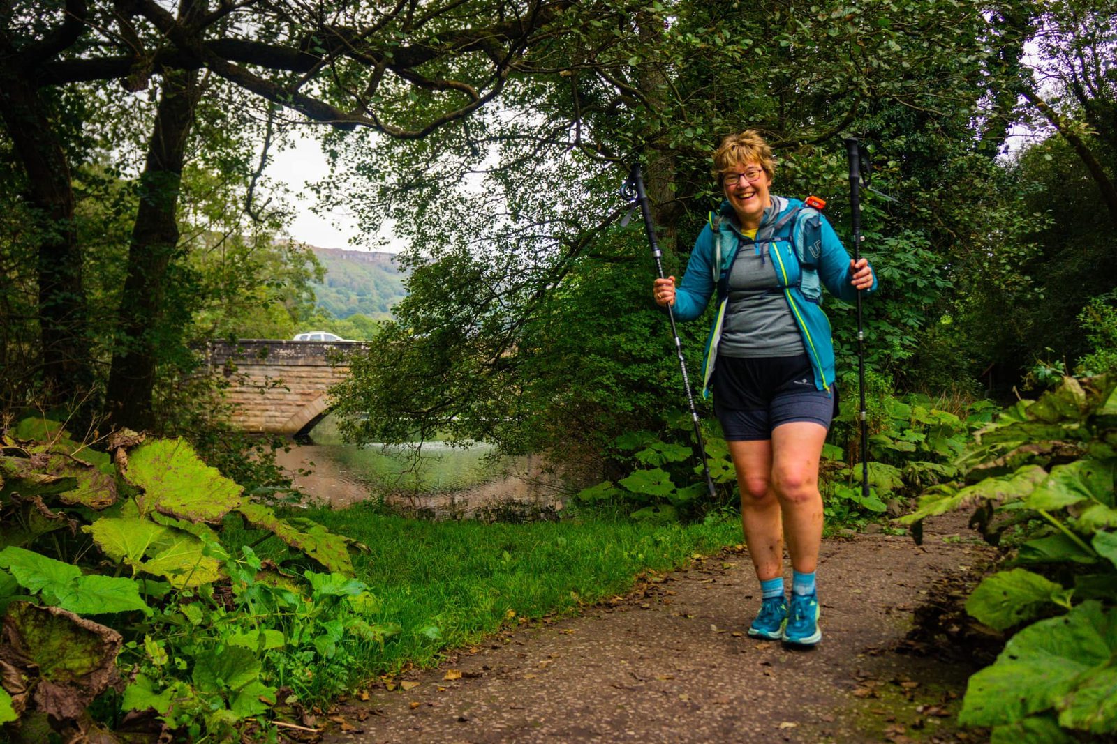 A person in outdoor attire and hiking gear, including a jacket and shorts, walks along a path surrounded by lush greenery. They are smiling, holding trekking poles, and a stone bridge can be seen in the background over a body of water.