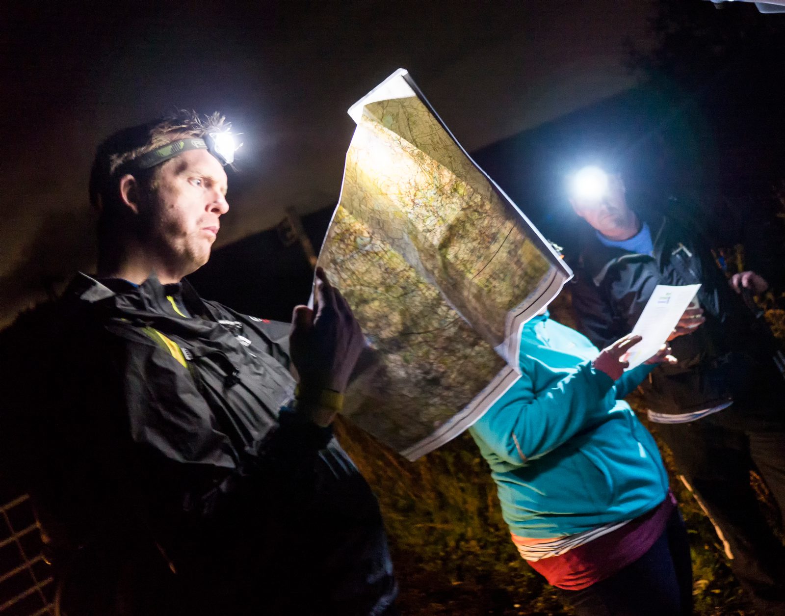 Three people wearing headlamps are looking at maps in a nighttime outdoor setting. The person in the foreground is holding a large map, while the other two are slightly obscured but also appear to be examining documents. The light from their headlamps illuminates the maps.