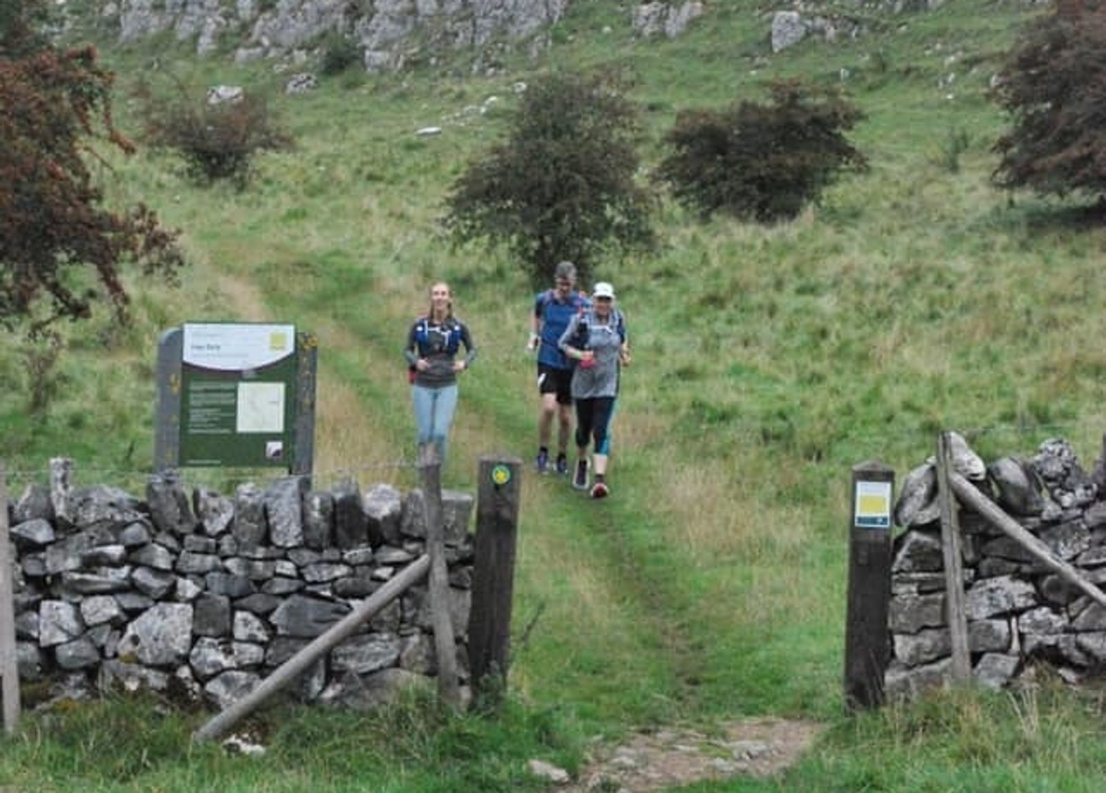 Three people jogging along a grassy trail in a rural, hilly area. They are approaching a stone gate with a wooden signpost on the left. Bushes and trees are scattered around; a rocky outcrop is visible in the background.