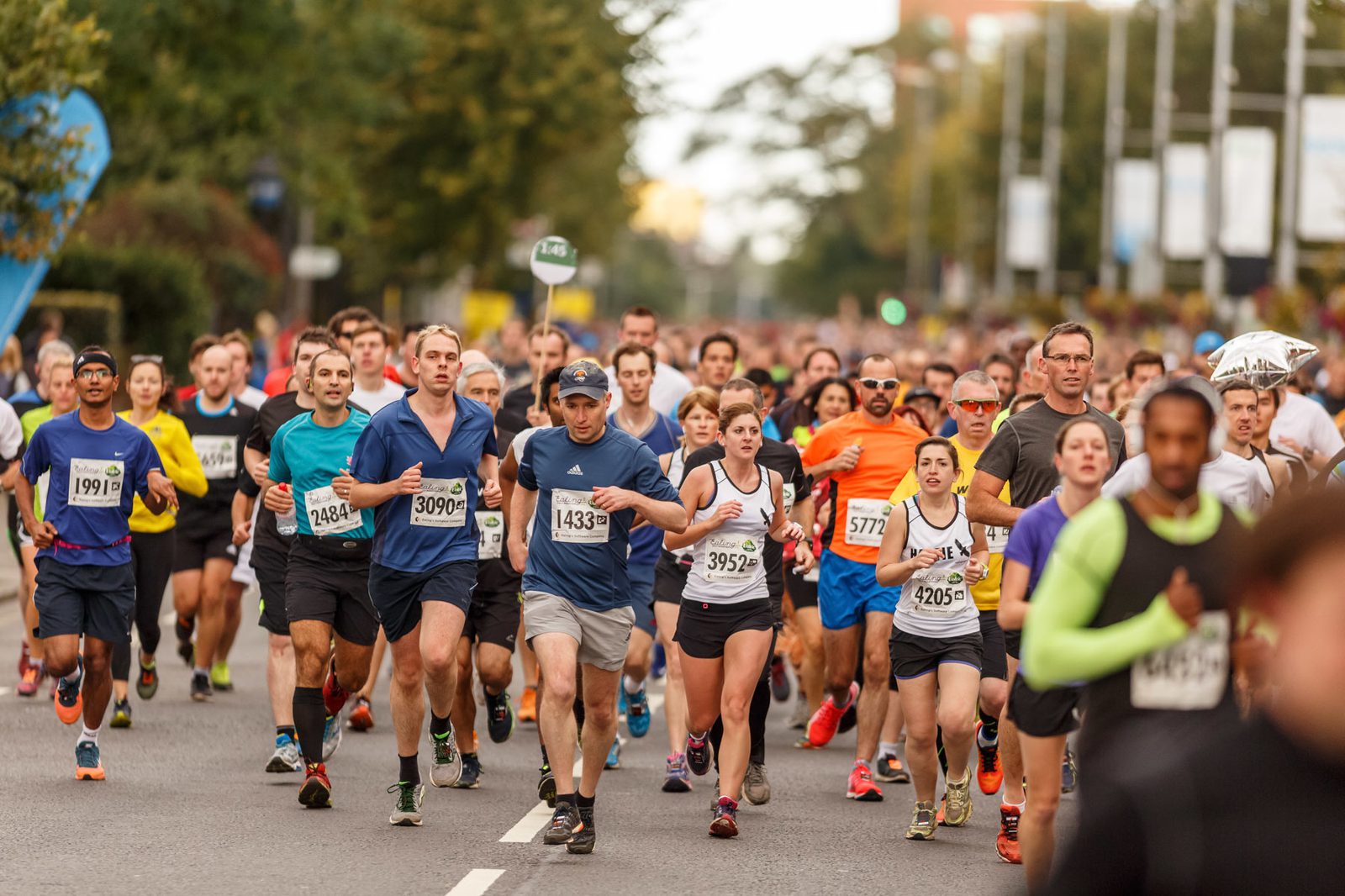 A large group of runners participating in a marathon. They are wearing race bibs with numbers and running on a road surrounded by trees. The runners display various expressions and are dressed in different athletic gear. The weather appears mild.