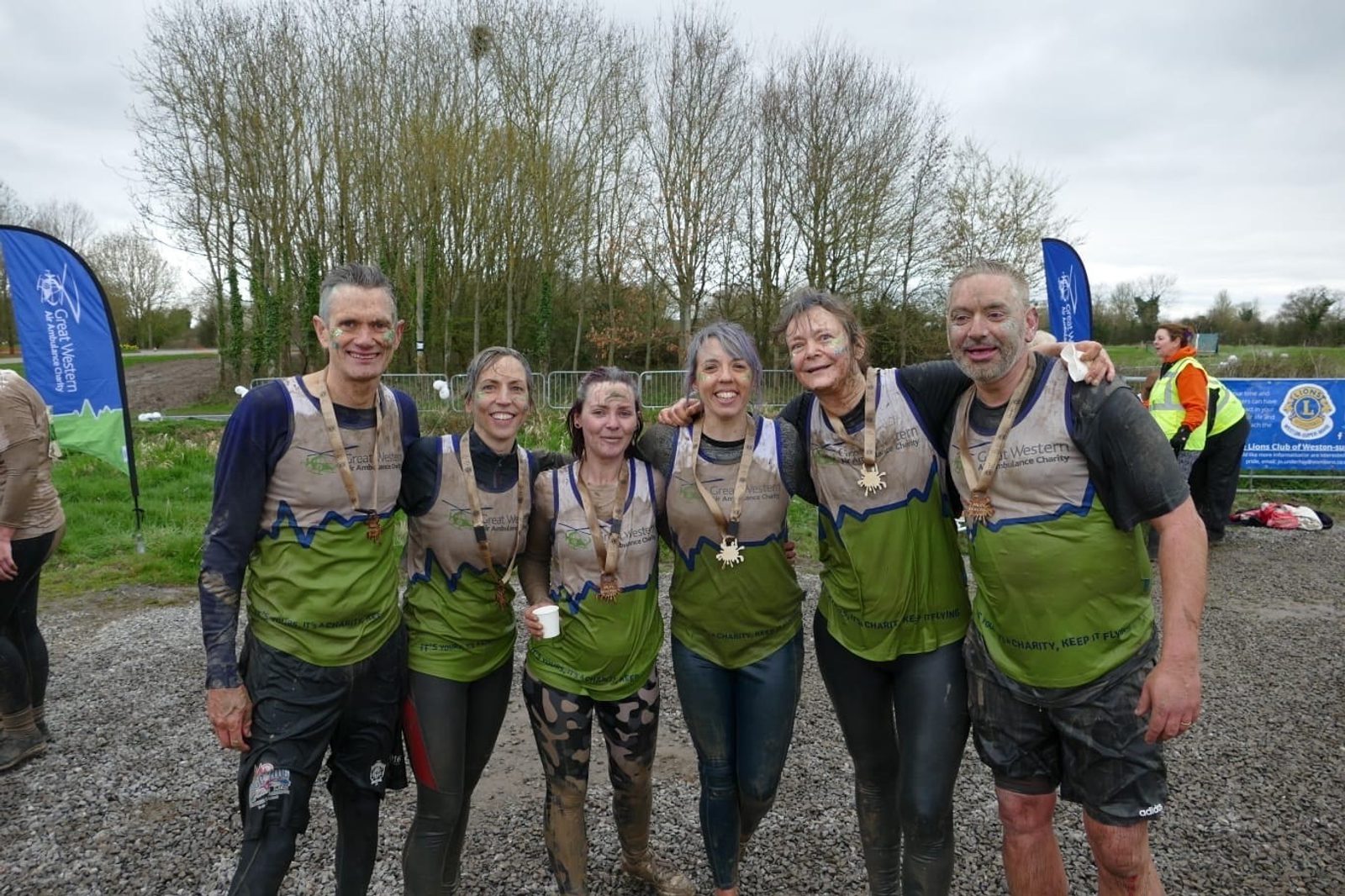 A group of six people, covered in mud, smile at the camera after completing an outdoor event. They stand closely together, wearing matching green shirts with medals around their necks. Trees and event banners are visible in the background.