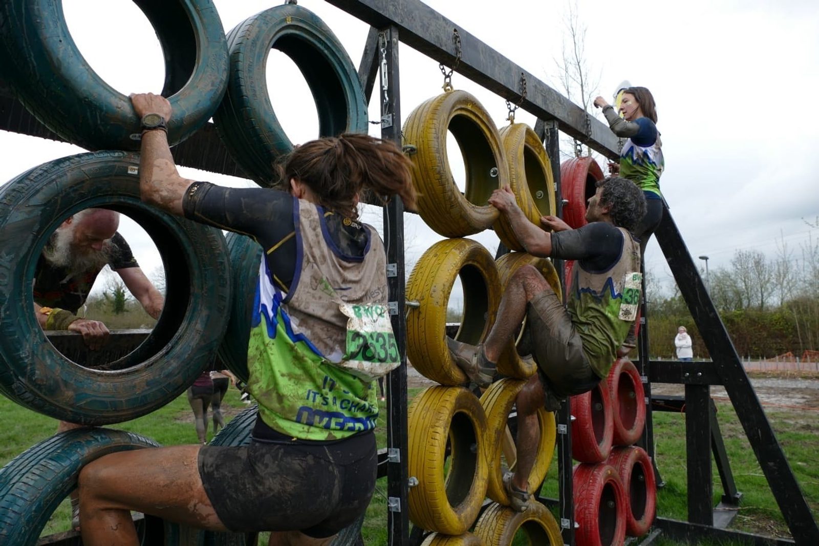 Participants in a muddy obstacle course race climb a wall made of colorful tires. They are wearing race bibs and are covered in mud. The atmosphere appears challenging and energetic as individuals strive to reach the top. Trees and a cloudy sky are in the background.
