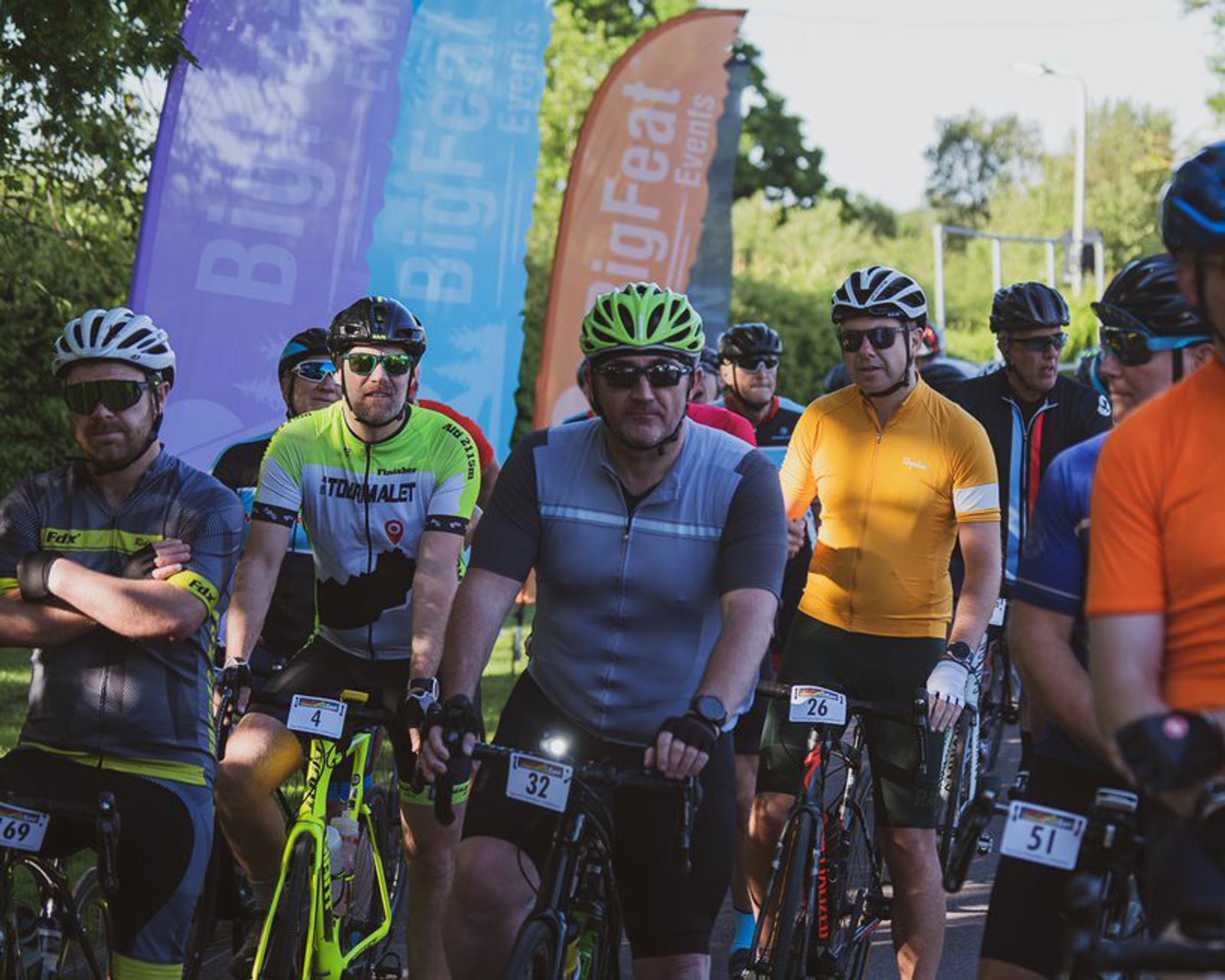 A group of cyclists wearing helmets and sunglasses are gathered at the starting line of a cycling event. Numbered tags are attached to their bikes and jerseys. Colorful banners for "Big Feat Events" are visible in the background, with trees lining the road.