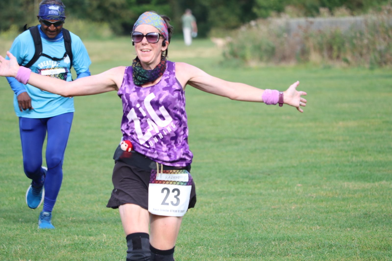 A woman wearing sunglasses, a colorful headscarf, a purple tank top, and a race bib numbered 23 is smiling with her arms outstretched while running on a grass field. A man in a blue outfit is running behind her. Trees and a trail are visible in the background.