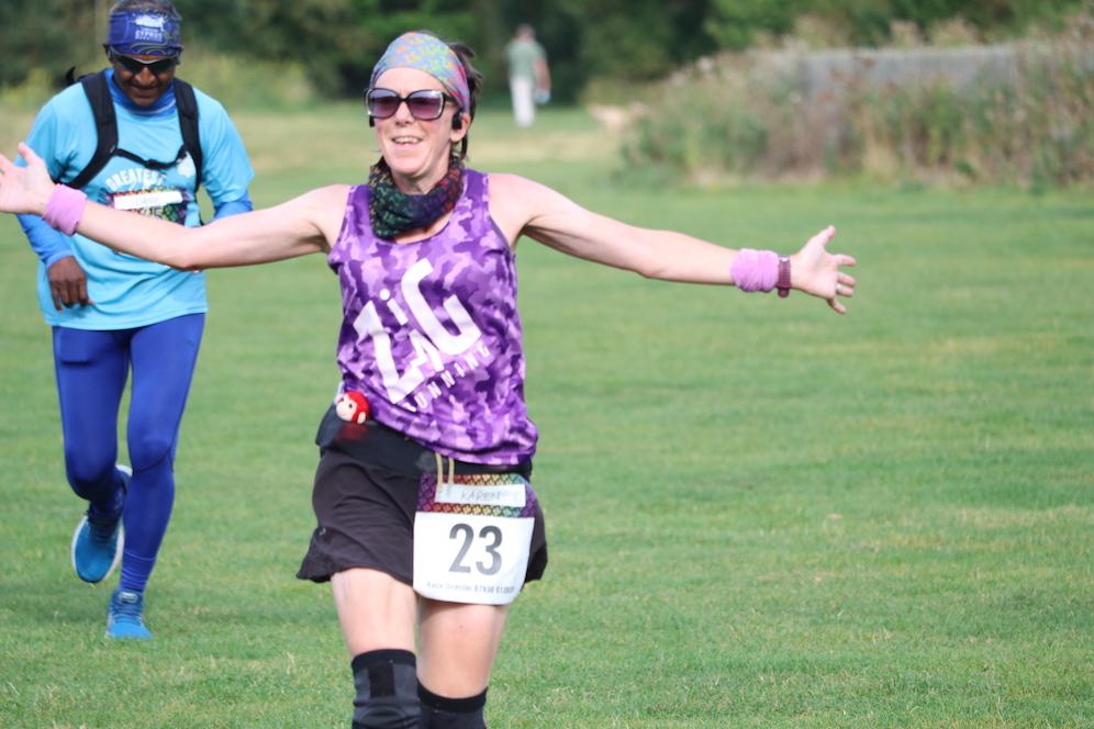 A woman wearing sunglasses, a colorful headscarf, a purple tank top, and a race bib numbered 23 is smiling with her arms outstretched while running on a grass field. A man in a blue outfit is running behind her. Trees and a trail are visible in the background.