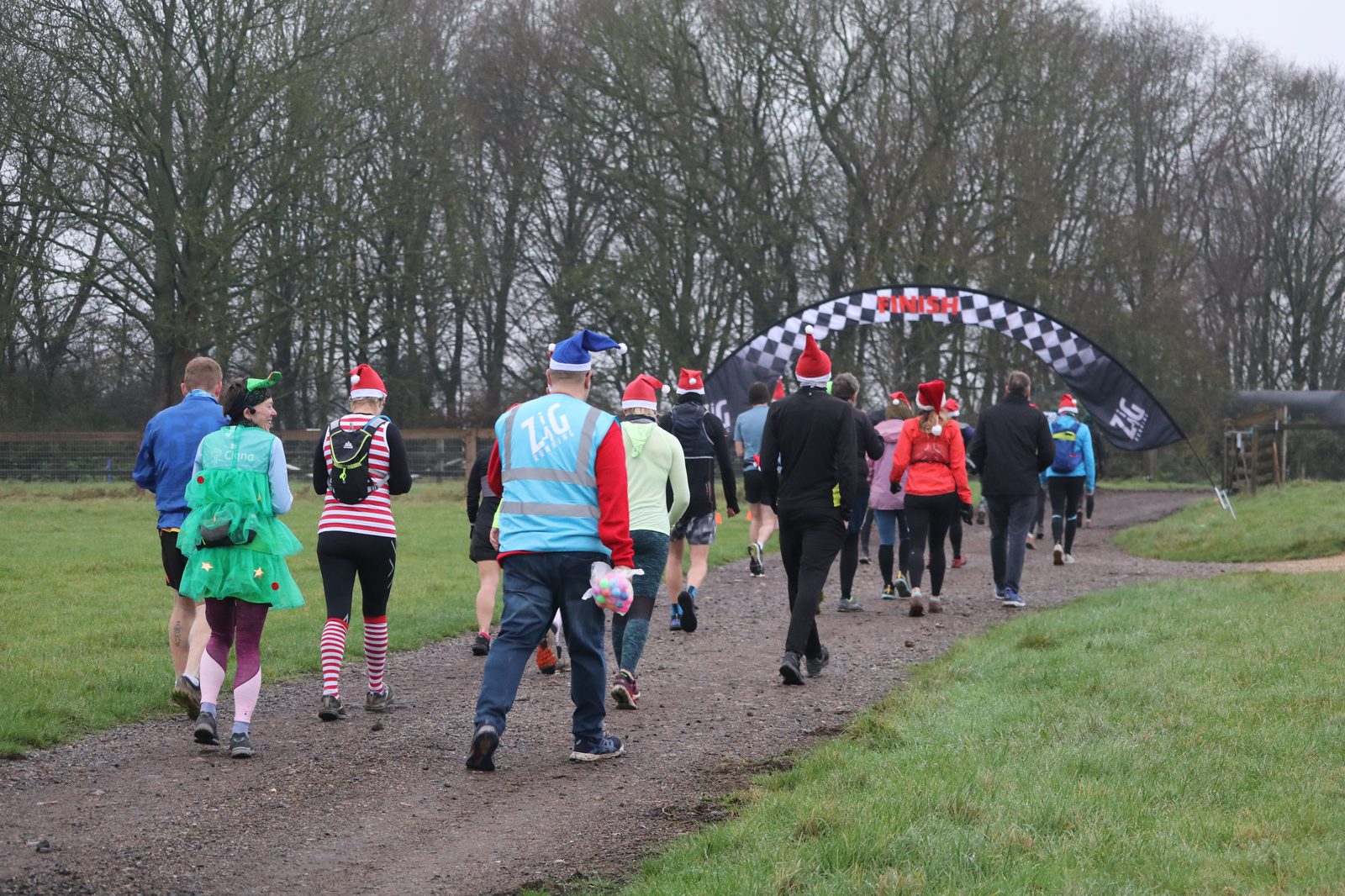 A group of people in festive clothing and hats are running on a gravel path towards a checkered finish line banner in a park setting. The runners are surrounded by green grass and trees on a cloudy day.