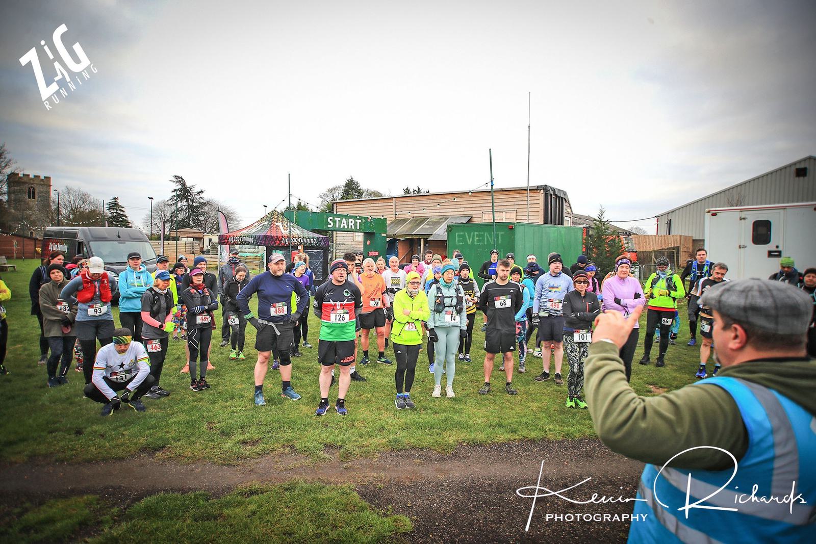 A large group of runners stands at the starting line of a race, listening to an organizer. They are dressed in a mix of athletic gear, with race bibs pinned to their clothes. The setting appears to be an outdoor event area with tents and essential amenities visible.