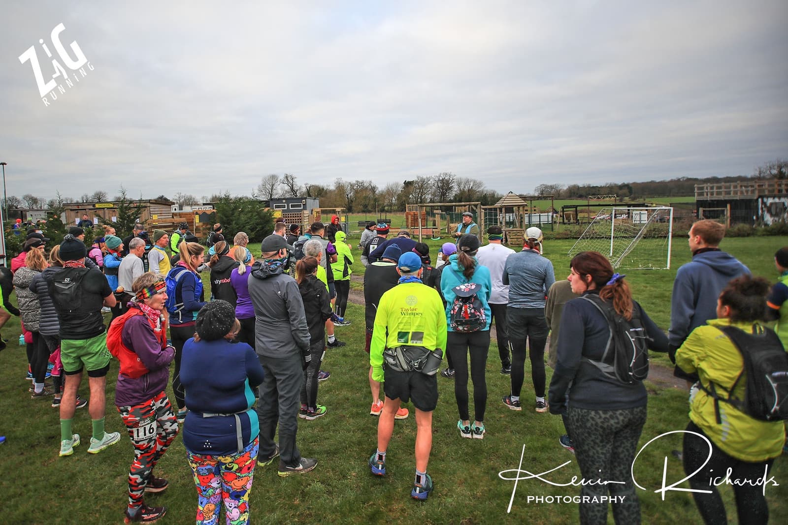A group of runners in colorful athletic wear gather on a grassy field, facing a speaker who appears to be giving instructions or speaking with a microphone. The background includes a soccer goal and several buildings. The sky is overcast.