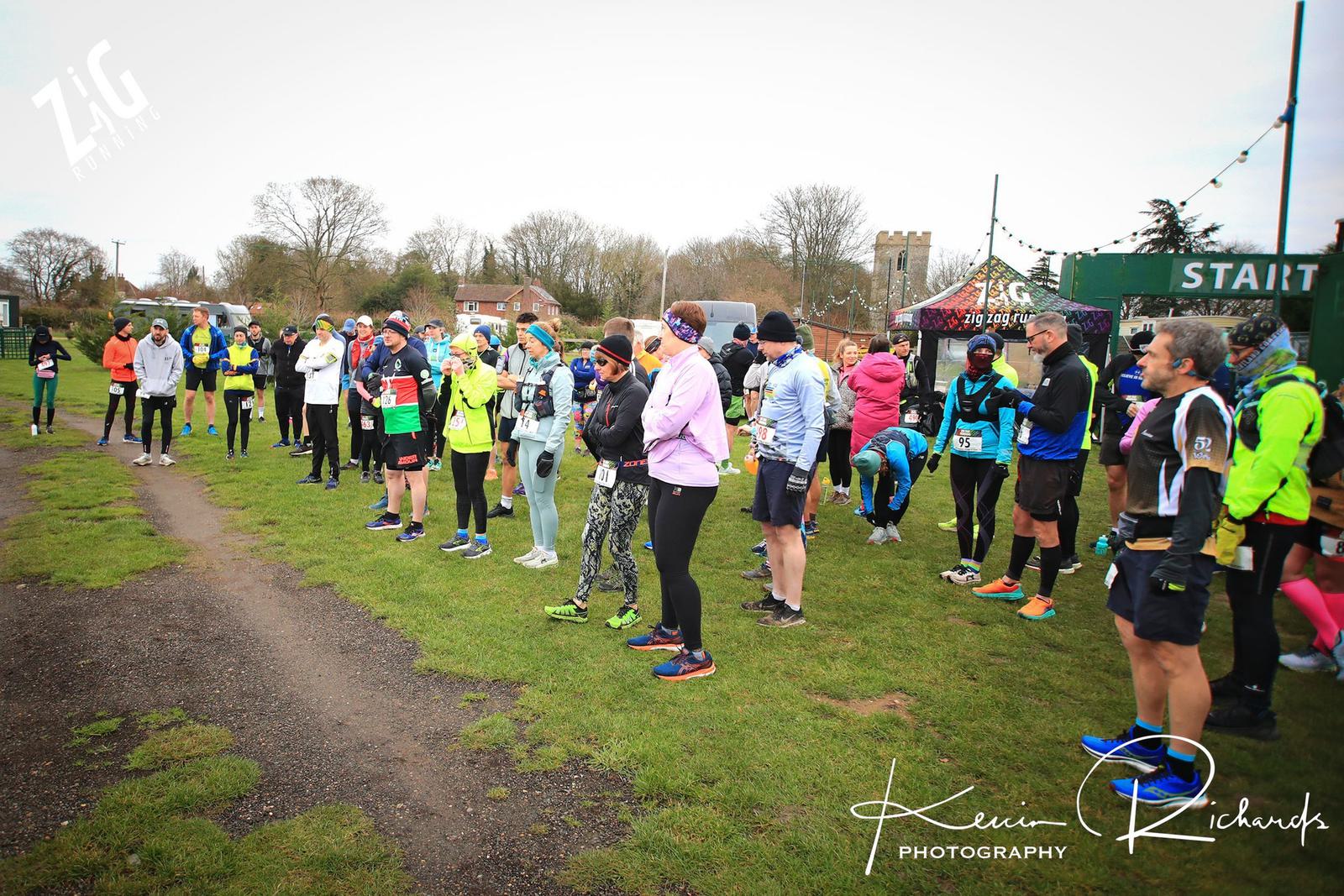 A group of runners gather near the starting line of a race on a grassy field. They are dressed in various running gear, some wearing bright colors. In the background, there are trees, a few buildings, and a church tower. A banner reading "START" is visible.