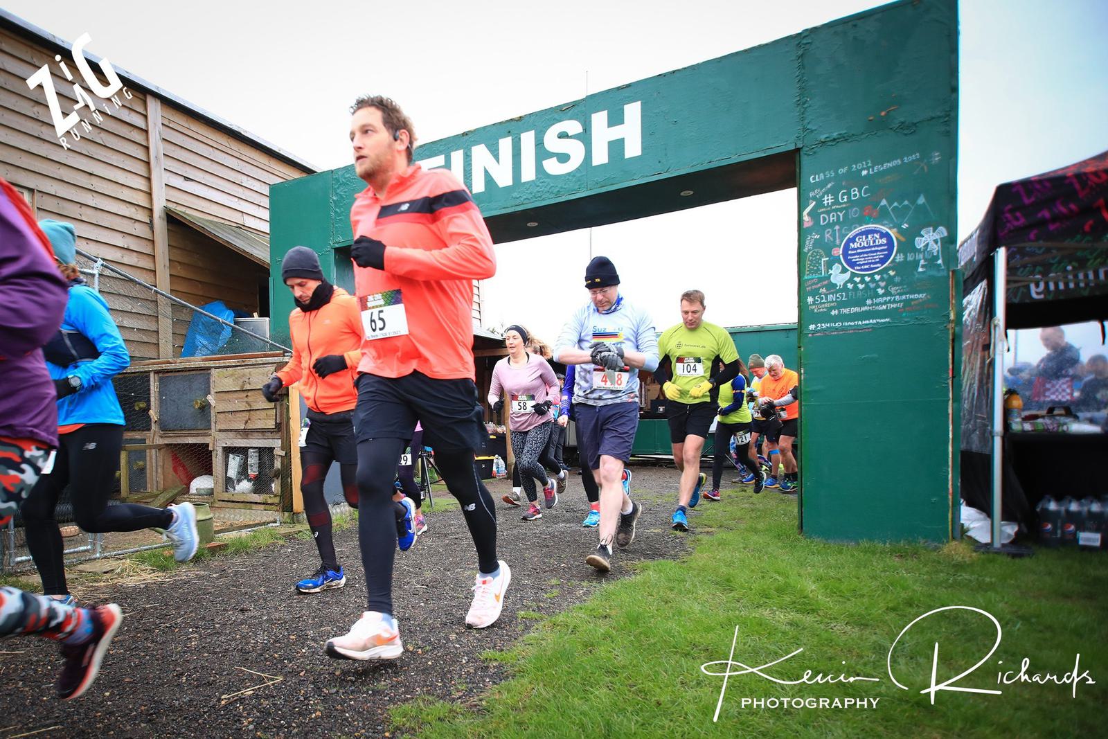 Runners crossing the finish line in an outdoor race. Participants are dressed in athletic gear, with some wearing bright colors. A green structure with "FINISH" written on it is visible in the background. The event appears to be taking place on a cloudy day.
