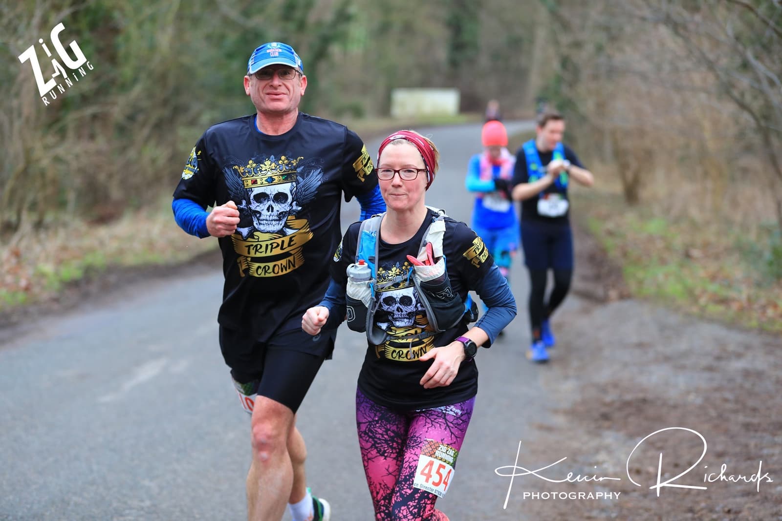 Two runners on a rural road participate in a race, wearing themed shirts featuring a skull with a crown and the words "Triple Crown." The male runner wears glasses and a hat, while the female runner wears a headband and carries water bottles. Other runners are visible behind them.