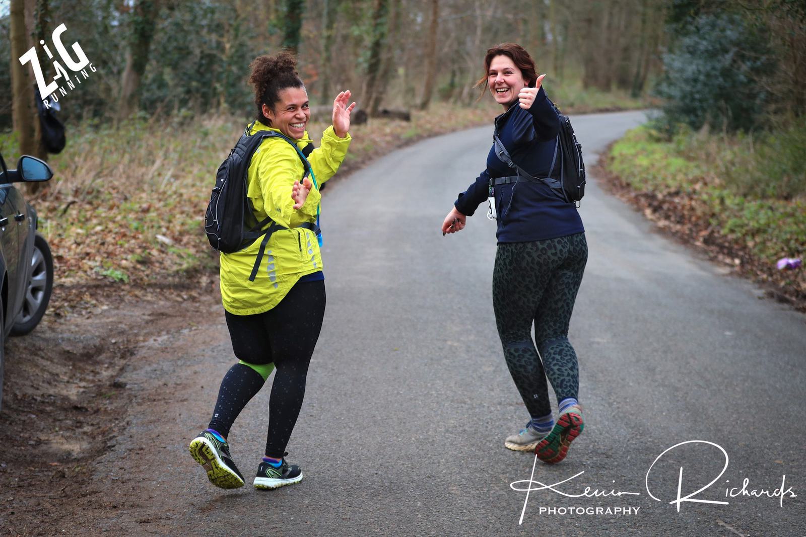 Two women wearing athletic clothing and backpacks joyfully run on a forested road, giving thumbs up. One wears a bright yellow jacket and the other dark clothing. The scene is lively and cheerful, with "Zig" and "Kevin Richards Photography" logos visible.