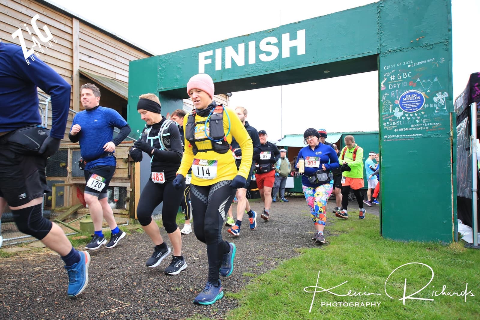 Several runners near a finish line, marked by a green archway reading "FINISH." The competitors, dressed in varying gear, briskly move forward, some with smiles. A chalkboard with writing and timing equipment is visible in the background. Kevin Richards Photography.