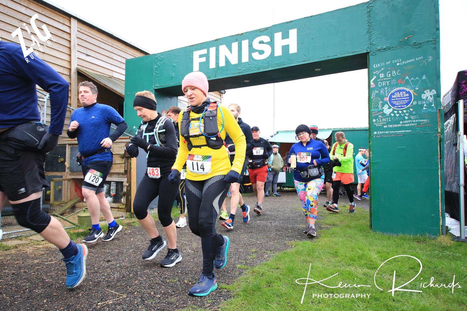 Runners crossing the finish line at a race, some looking exhausted while others appear energized. The backdrop features a green finish banner and nearby buildings. They wear numbers pinned to their shirts, ranging from 114 to 143, with various colored outfits and gear.