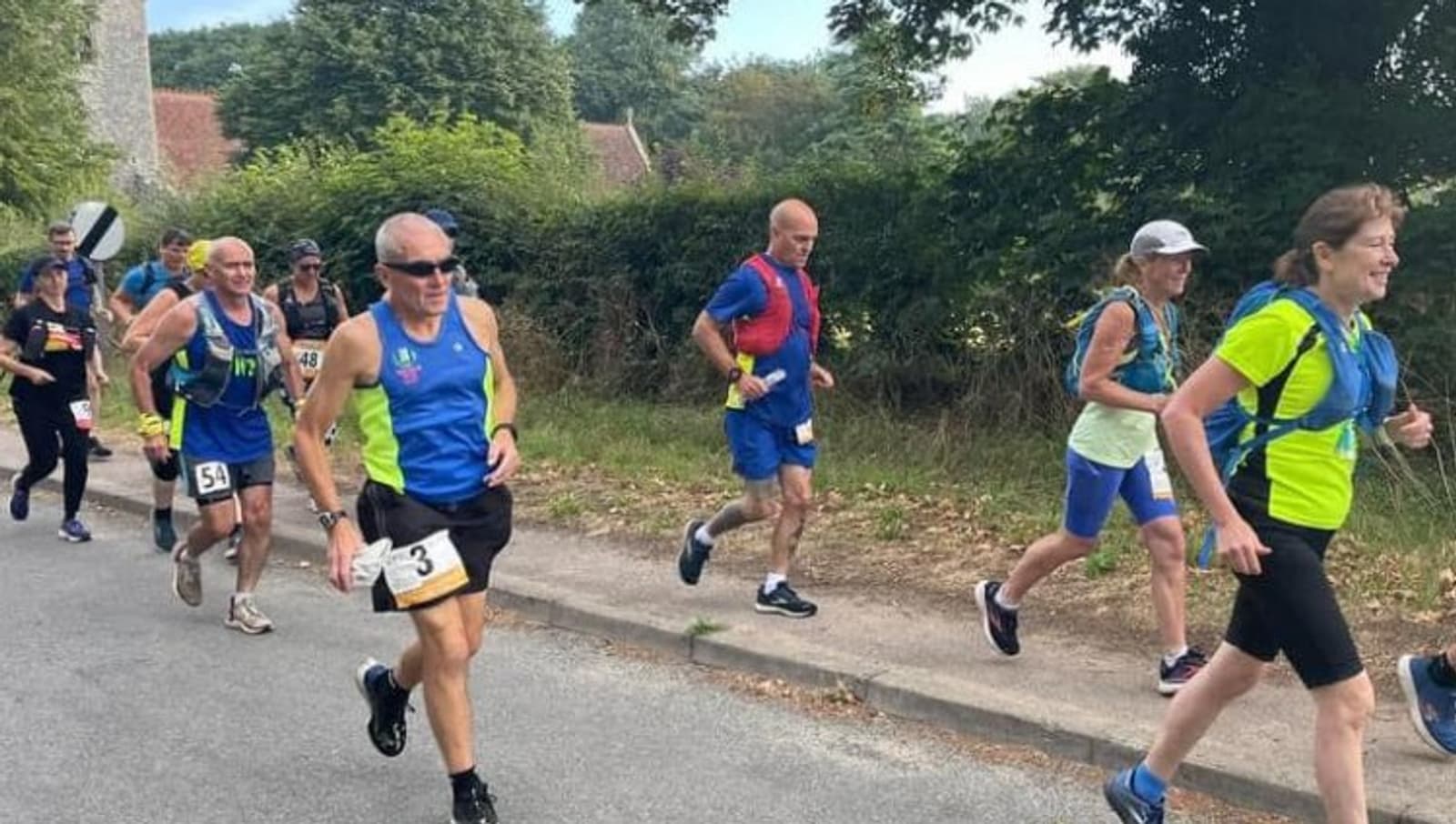 A group of marathon runners jogs along a roadside surrounded by greenery. Some runners wear blue outfits, while others are in different colored athletic gear. The atmosphere appears to be sunny and bright, with participants looking focused and determined.