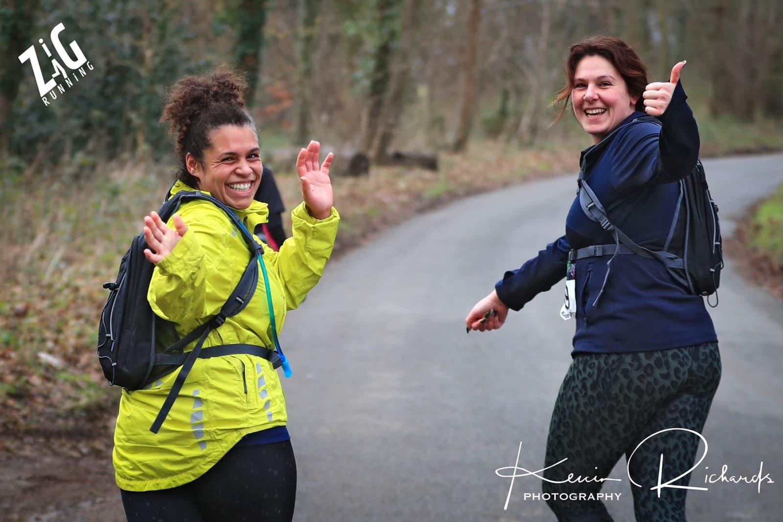 Two people on a wooded path smile and give thumbs up to the camera while wearing running gear. One wears a bright yellow jacket and the other a dark jacket with leopard-patterned leggings. Trees and a winding road are visible in the background.