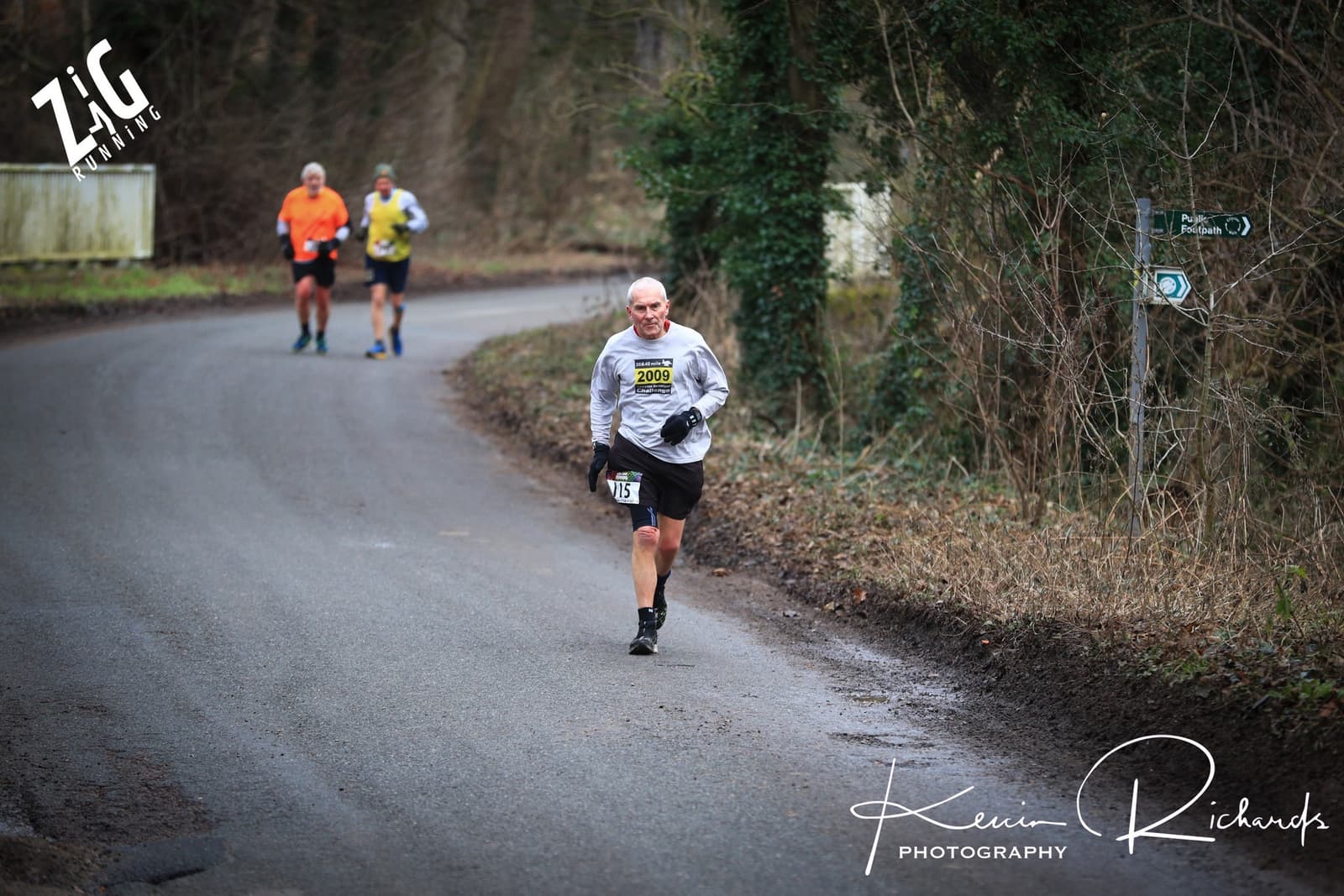 A group of runners participates in a road race in a forested area. The lead runner, wearing a gray shirt and black shorts with the number 2000, appears focused. Two runners are behind him. Directional race signs are seen along the trail. Photo credit: Kevin Richards Photography.