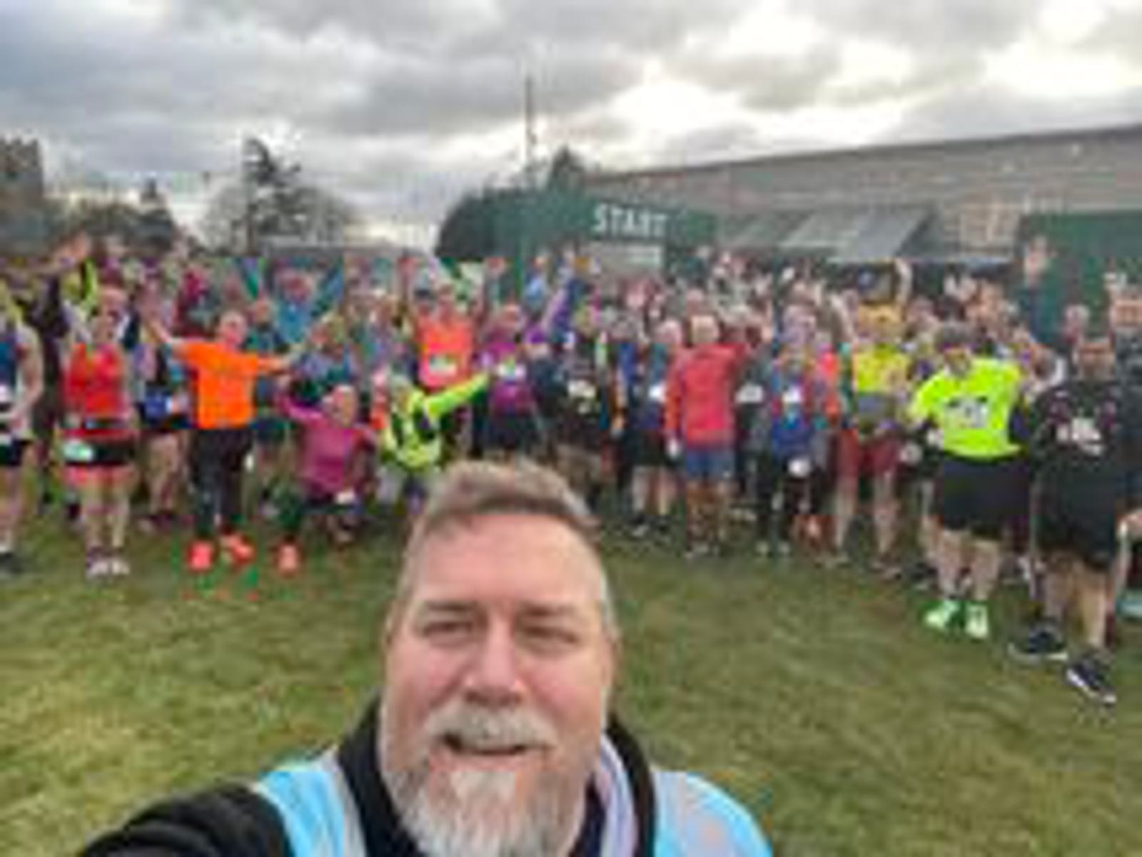 A bearded man takes a selfie in front of a large group of runners standing on grass. The runners are wearing brightly colored athletic gear, and many have their arms raised. A green "START" banner is visible in the background. The sky is overcast.