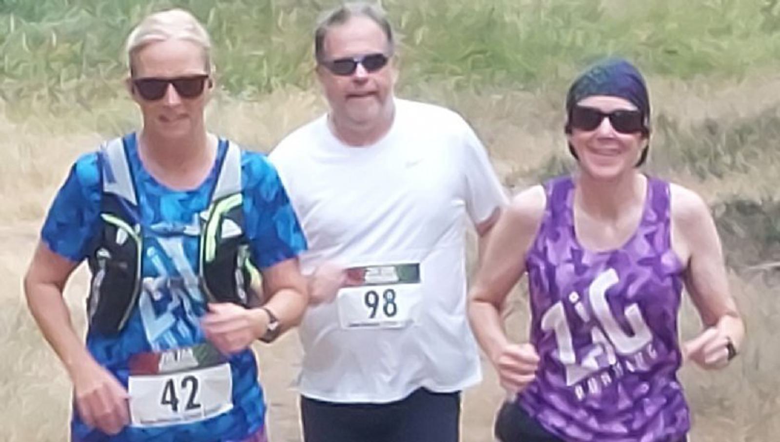 Three runners participate in an outdoor race. Two women and one man run side by side, with bib numbers 42, 98, and 1AG on their shirts. The woman on the left wears a blue shirt with a hydration pack, the man wears a white shirt, and the woman on the right wears a purple shirt.