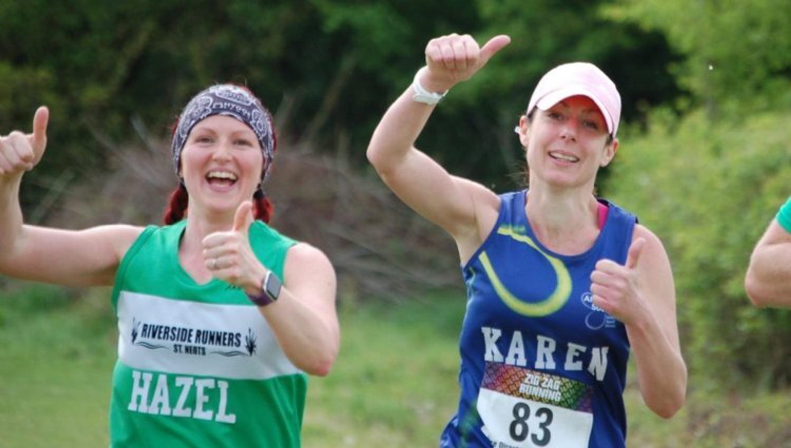 Two women running outdoors and giving thumbs-up signs. The woman on the left is wearing a green "Riverside Runners" shirt and a headband labeled "Hazel." The woman on the right is wearing a blue shirt numbered 83 and labeled "Karen." Trees and greenery are visible in the background.