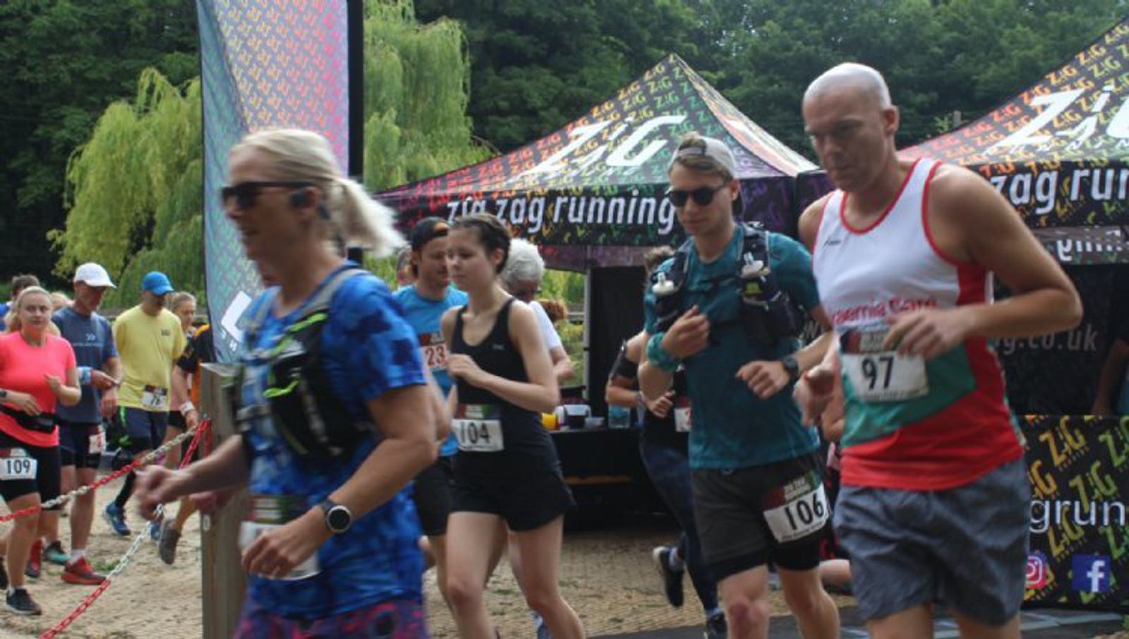 A group of people participating in a running race pass by a tent with "zig zag running" branding. Some wear bib numbers and hydration packs, and the background shows trees and more race-related tents. Everyone appears focused and in motion.