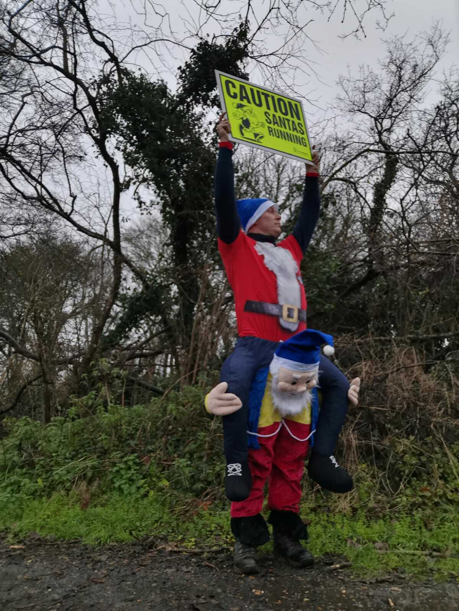 A person dressed in a Santa Claus costume appears to be riding on the shoulders of a large puppet Santa Claus, holding a bright yellow sign that reads "CAUTION SANTAS RUNNING." They are outdoors near some leafless trees and bushes.