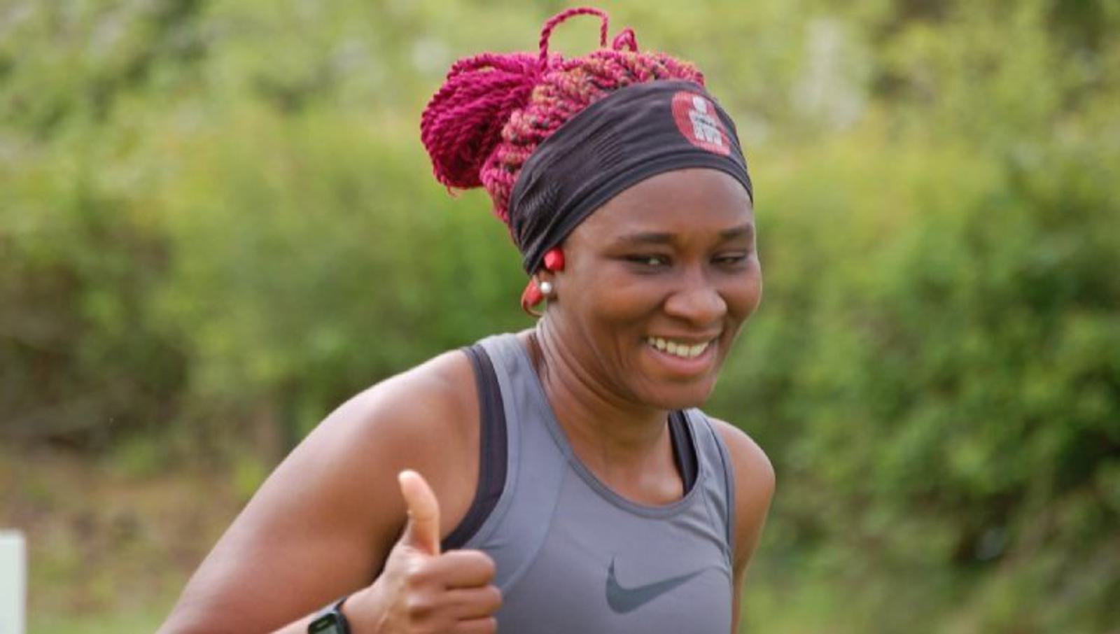 A woman with pink braided hair, wearing a black headband and gray sleeveless top, smiles and gives a thumbs-up while jogging outdoors. There are green trees in the background.
