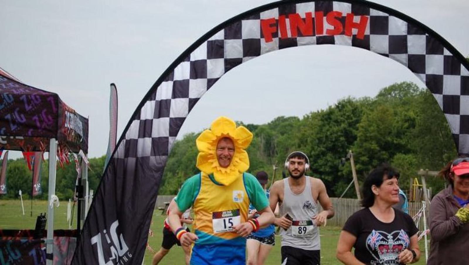 Participants are nearing the finish line of a race. One runner in a flower costume with number 15 stands out, smiling broadly. Others, dressed in typical running gear, follow closely behind. The finish line arch is checkered with "FINISH" in red at the top.