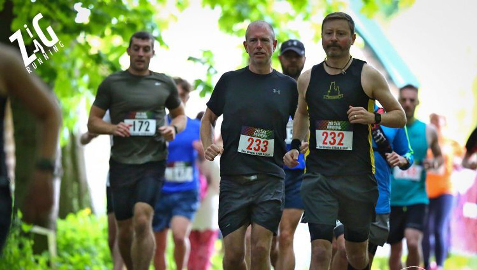 A group of runners participating in a race on a sunny day, surrounded by greenery. The runner in the foreground, wearing black and numbered 233, is running alongside another person wearing black with yellow accents and numbered 232. A crowd follows behind them.