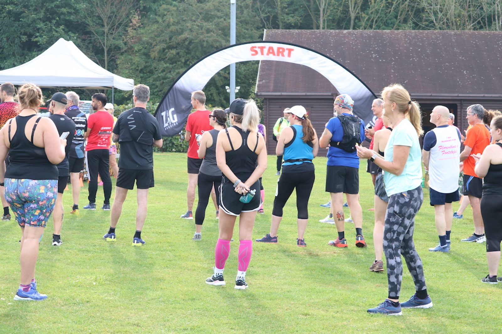 A group of people in athletic clothing gather near a starting line, marked by a white and black banner with "START" written in red. They appear to be preparing for a race or run. Trees and buildings are visible in the background.