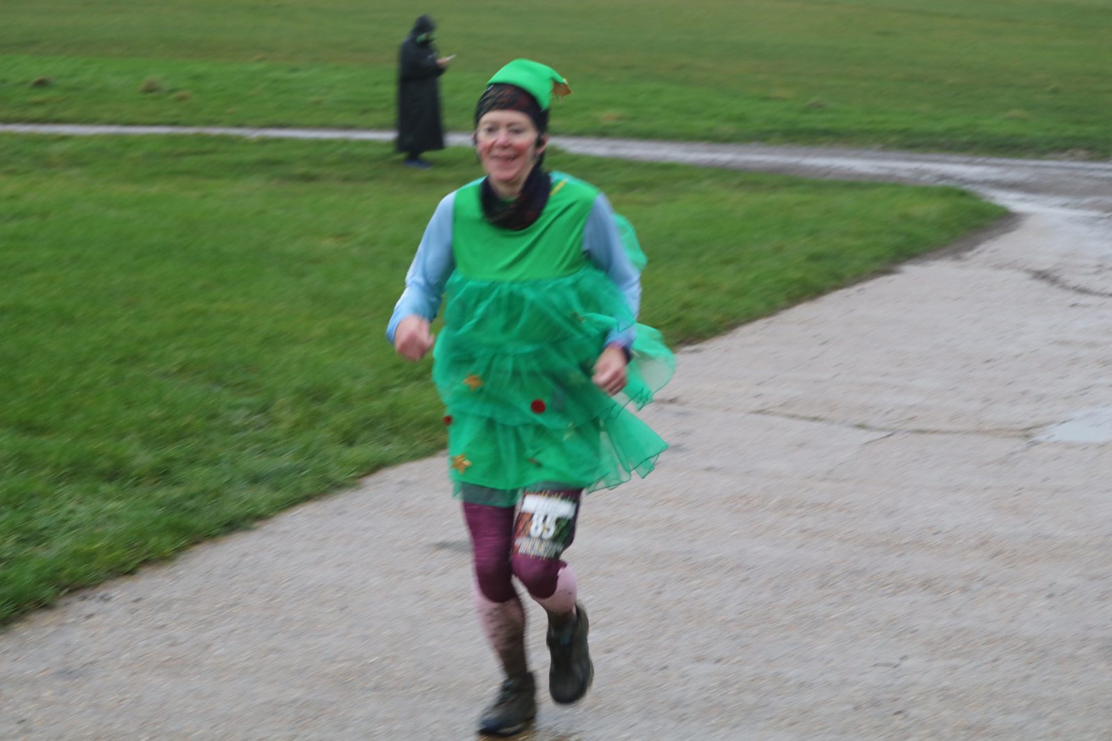 A person runs outside on a path, dressed in a green Christmas tree costume with red ornaments. They are smiling and wearing running shoes with a race number attached. The background is grassy, and another person in dark clothing is seen in the distance.