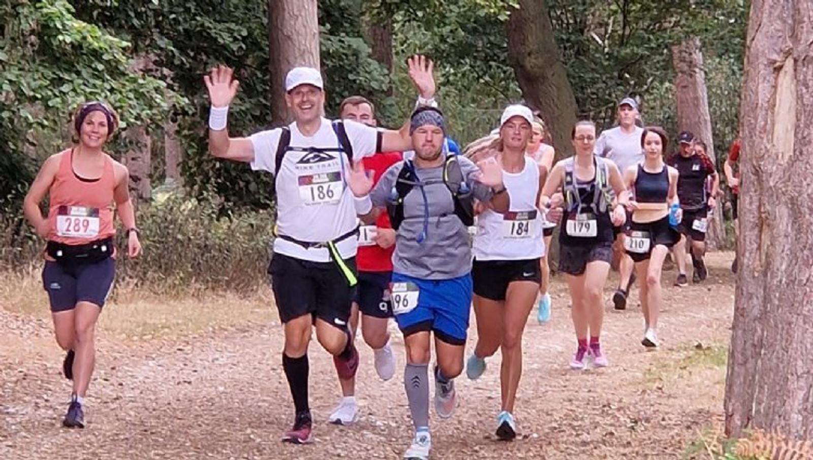 A group of runners on a forest trail during a race. The leading runner, wearing a white shirt and black shorts with number 186, has both hands raised in the air. Other runners, with numbers 181, 208, and 179, follow closely behind. Trees and foliage surround the path.