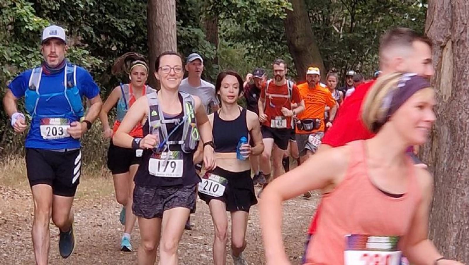 A group of runners participating in a trail race. They are jogging on a dirt path surrounded by trees. The runners wear race bibs and various athletic outfits, including tank tops, shorts, and hydration packs. The mood appears focused and determined.