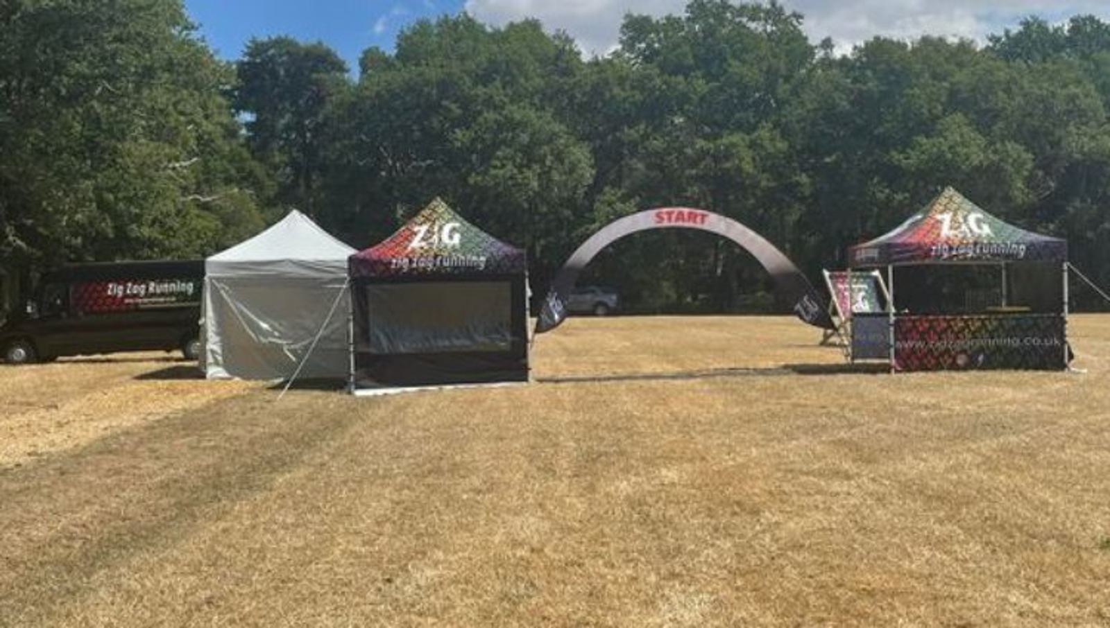 A grassy field with two event tents, a black van, and a START banner in the center. The left tent is white, and the right tent has "Zig Zag Running" branding. In the background, there are trees and a partly cloudy sky.