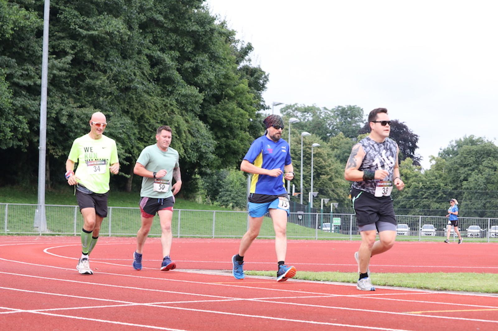 Four men jog on a red running track, each wearing different colored athletic clothing and numbered bibs. In the background, there are trees, fences, and a parked car. The sky is overcast, and another runner can be seen in the distance.