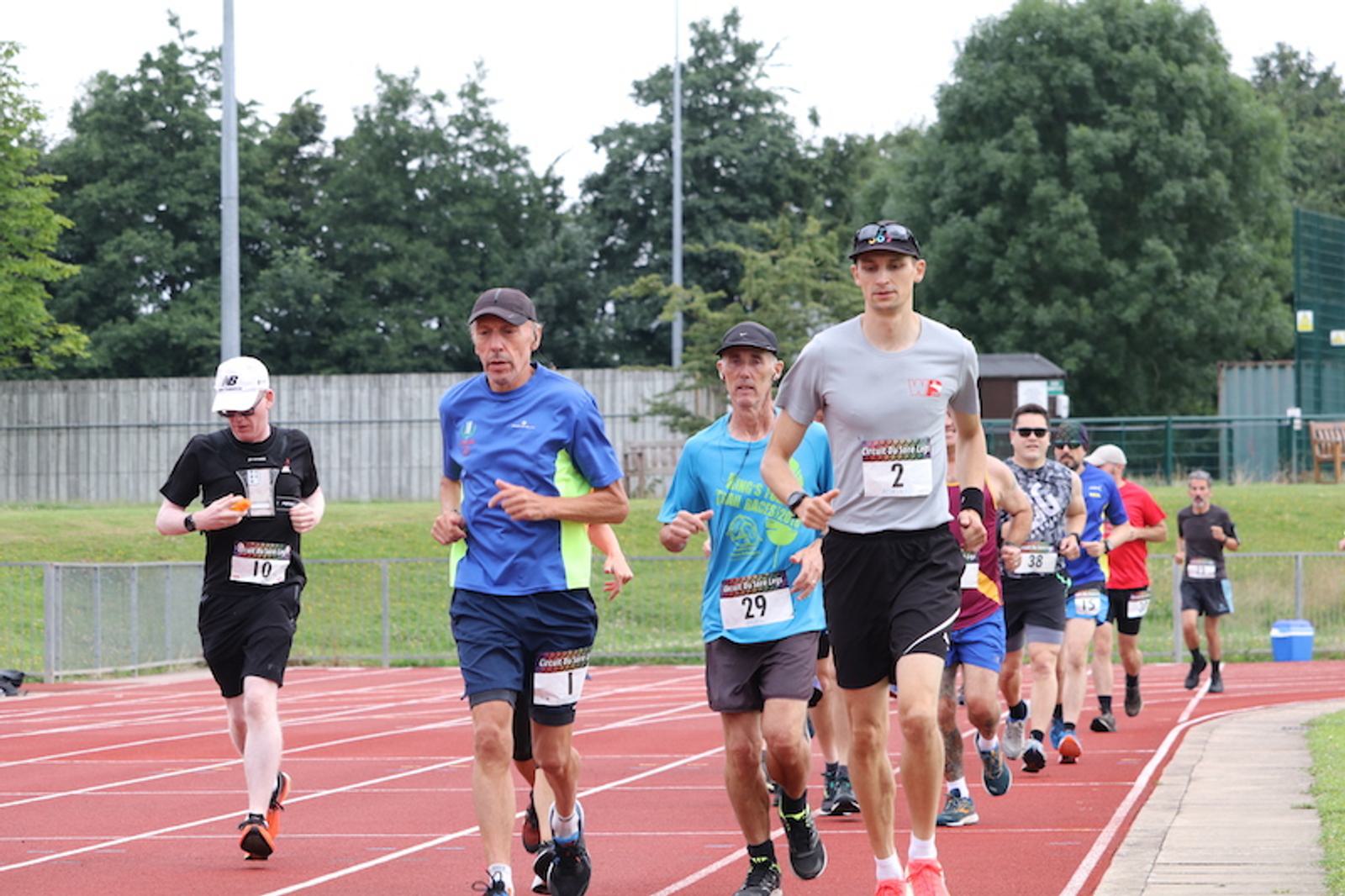 A group of runners on an outdoor track, wearing race bibs and athletic wear. The runners appear focused, each in mid-stride. The background includes trees, a fence, and a grassy area, indicating a park or sports facility. The sky is clear, suggesting a sunny day.